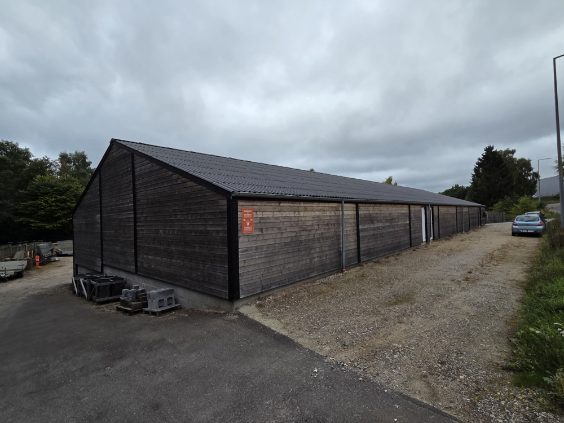 Mørk træbeklædt erhvervshal set fra gavlen, med grusvej langs siden