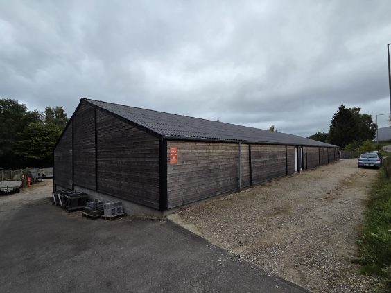 Mørk træbeklædt erhvervshal set skråt fra gavlen, med grusvej og en parkeret bil langs langsiden